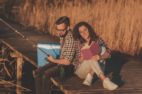 Couple sitting at the lake docks, man fishing while woman is reading a book, enjoying a peaceful day in nature and relaxing