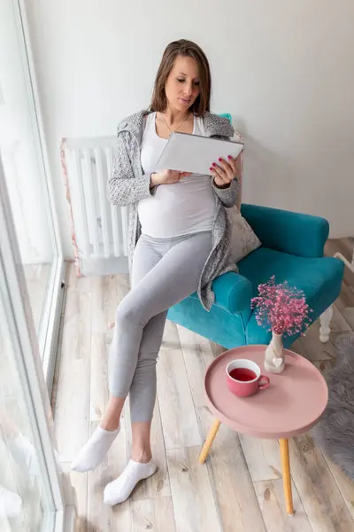 自宅でリラックスした美しい妊婦 アームチェアに座ってタブレットコンピュータで産休ブログを読む — ストック写真