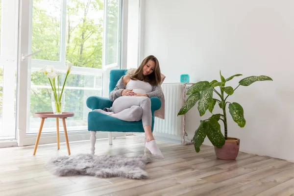 自宅でリラックスして赤ちゃんを期待する美しい若い妊娠中の女性 椅子に座っている妊婦 彼女の腹と笑顔を保持 — ストック写真