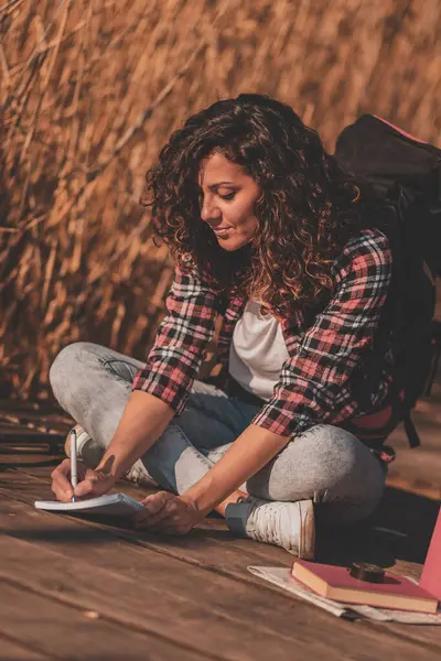 Young female scientist wearing a backpack sitting on wooden lake docks on a sunny autumn day, taking notes while on field work trip