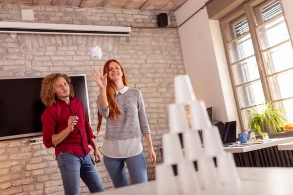 Business people having fun at the office, playing office games while on a break, tearing down a pyramid of plastic cups with a paper ball