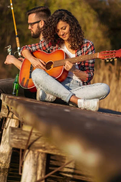 Göl rıhtımında oturan bir çift, erkek balık tutarken kadın gitar çalıyor, doğada huzurlu bir günün tadını çıkarıyor ve dinleniyor.