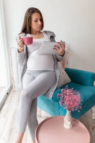 自宅でリラックスした美しい妊婦 椅子に座って紅茶を飲み タブレットコンピュータでネットサーフィン — ストック写真