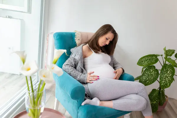 自宅でリラックスして赤ちゃんを期待する美しい若い妊娠中の女性 椅子に座っている妊婦 彼女の腹と笑顔を保持 — ストック写真