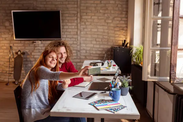 İki iş arkadaşı ofiste, akıllı bir telefonla selfie çekiyorlar; serbest çalışanlar birlikte çalışıyorlar ve tatilde eğleniyorlar.