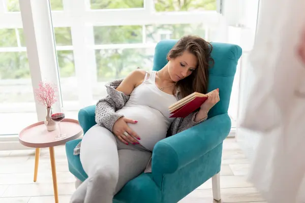 窓からアームチェアに座り 本を読み ブラックベリーワインを飲む美しい妊婦 — ストック写真