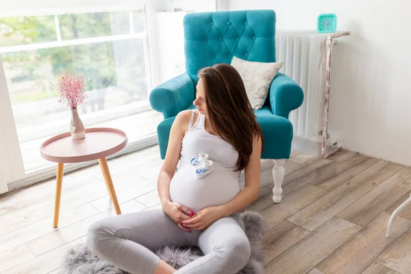 自宅でリラックスし 赤ちゃんを期待して 美しい妊娠中の女性 アームチェアの隣に座っている妊婦 彼女の腹に小さな赤ちゃんの靴を抱き — ストック写真