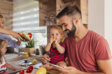 Genç baba oğlunu kucağında tutuyor ve karısıyla ve kızıyla yemek pişirirken tablet bilgisayar kullanarak yemek tarifleri arıyor.
