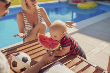 Ebeveynler sıcak güneşli yaz günlerini yüzme havuzunun yanında geçiriyorlar. Oğullarıyla, babaları da oğlunu bir dilim karpuzla besliyor.