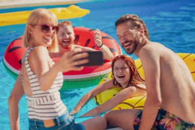 Güneşli bir yaz gününde yüzme havuzunda eğlenen iki güzel genç çift, akıllı telefon kullanarak selfie çekiyorlar, dinleniyorlar ve havuz başında güneşleniyorlar.
