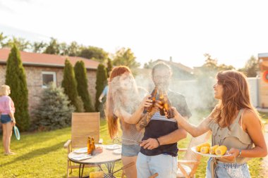 Bir grup neşeli genç arkadaş arka bahçedeki barbekü partisinde eğleniyor, et pişirirken bira şişeleriyle kadeh kaldırıyor, güneşli bir yaz gününde dinleniyor.