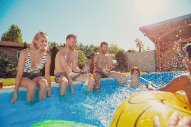 Bir grup neşeli genç arkadaş açık hava yaz partisinde eğleniyor, havuz kenarında oturuyor, bira içiyor ve birbirlerine su sıçratıyorlar.
