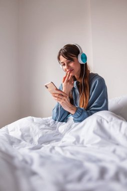 Pijama giyen, sabahları evde dinlenen, kulaklık takan, müzik dinleyen ve akıllı telefon kullanan güzel genç bir kadın.