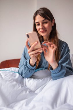 Güzel genç bir kadın pijama giyiyor ve sabah uyandıktan sonra yatakta akıllı telefon kullanıyor.