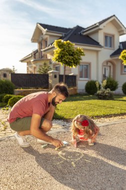 Baba ve kızı evlerinin önünde renkli tebeşirlerle resim çizerken eğleniyorlar.