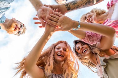 Bir grup neşeli gencin düşük açılı görüntüsü bir araya toplanmış, elleri ortada birleşmiş kameraya bakıyordu.