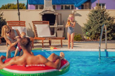 Neşeli ebeveynler yüzme havuzunun kenarında çocuklarıyla oynayıp birbirlerine su sıçratarak yaz tatilinin tadını çıkarıyorlar.