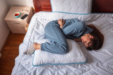 Genç kadın ağrı içinde yatakta kıvrılmış yatıyor, regl döneminde karnı ağrıyor; depresif yalnız kadın sabahları yatakta yatıyor