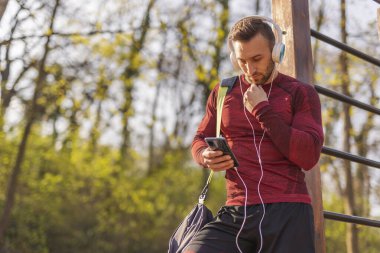 Spor çantası taşıyan, kulaklık takan ve akıllı telefon kullanan aktif genç bir adam. Açık hava egzersizinden sonra mesaj yazıyor.