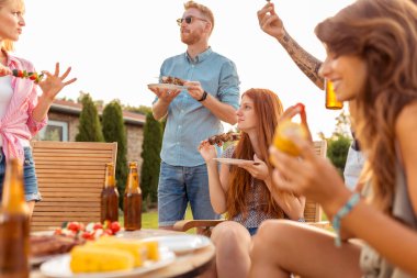 Bir grup neşeli genç arkadaş masanın etrafında toplanmış ızgara et yiyor, bira içiyor ve arka bahçedeki barbekü partisinde eğleniyorlardı.