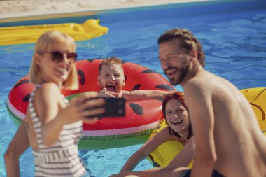 Güneşli bir yaz gününde yüzme havuzunda eğlenen iki güzel genç çift, akıllı telefon kullanarak selfie çekiyorlar, dinleniyorlar ve havuz başında güneşleniyorlar.