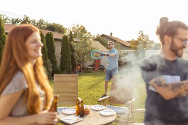Bir grup neşeli genç arkadaş arka bahçede barbekü partisi veriyor, et pişiriyor, bira içiyor, badminton oynuyor ve güneşli bir yaz gününde dışarıda dinleniyor.