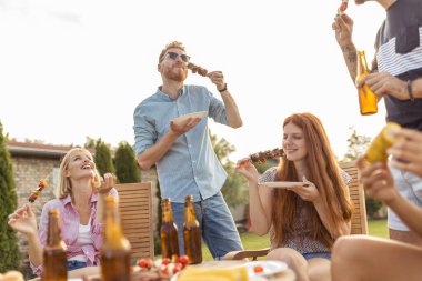 Bir grup neşeli genç arkadaş masanın etrafında toplanmış ızgara et yiyor, bira içiyor ve arka bahçedeki barbekü partisinde eğleniyorlardı.