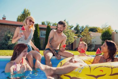 Bir grup genç arkadaş yaz havuz partisinde eğleniyor, havuz kenarında oturuyor ve karpuzlu dondurma yiyor, güneşli yaz günlerini dışarıda geçiriyorlar.