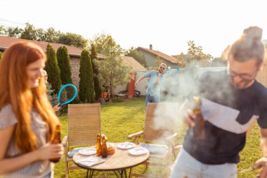 Bir grup neşeli genç arkadaş arka bahçede barbekü partisi veriyor, et pişiriyor, bira içiyor, badminton oynuyor ve güneşli bir yaz gününde dışarıda dinleniyor.