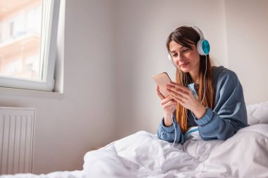 Pijama giyen, sabahları evde dinlenen, kulaklık takan, akıllı telefon kullanarak internette sörf yapan güzel bir genç kadın.