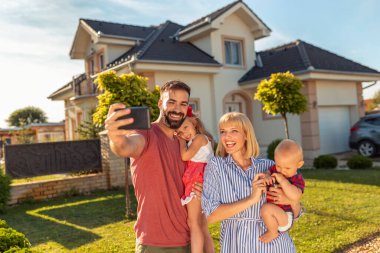 Güzel mutlu aile yeni evlerinin önünde akıllı telefonları kullanarak selfie çekerken eğleniyor.
