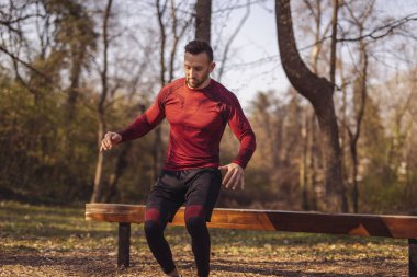 Aktif bir genç adam güneşli bir sonbahar gününde bir sokak idman parkında çalışıyor, bankın üzerinden atlıyor.