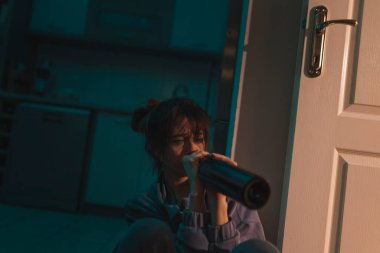 Üzgün sarhoş bir kadın karanlıkta yerde makyajla oturmuş şişeden şarap içiyor ve ağlıyor.