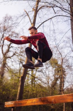 Atletik genç adam güneşli bir bahar gününde bir sokak idman parkında egzersiz yapıyor, bankın üzerinden atlıyor.
