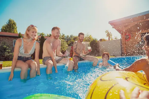陽気な若い友人のグループは 屋外の夏のパーティーで楽しんで プールに座って ビールを飲み お互いに水を飛ばしています — ストック写真