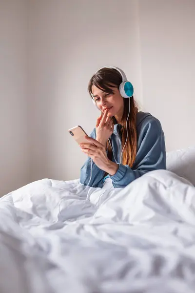 Beautiful young woman wearing pajamas sitting in bed, relaxing at home in the morning, wearing headset listening to the music and using smart phone
