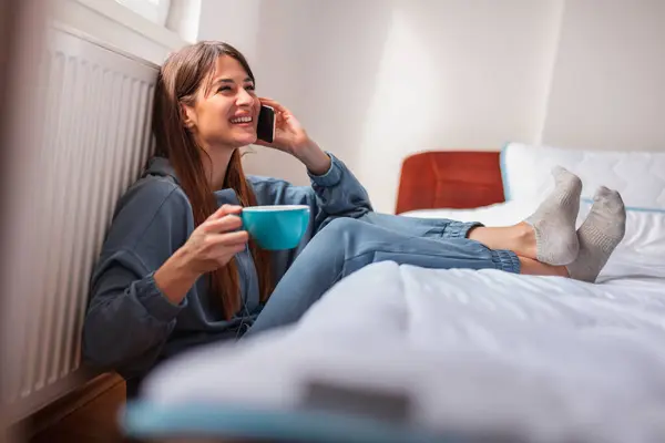 Beautiful young woman wearing pajamas sitting on the floor by the bed in bedroom, drinking coffee and having phone conversation