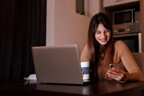 フリーランサーが自宅のオフィスで机に座り コーヒーを飲んで夜遅く働いている間にスマートフォンを使用する — ストック写真