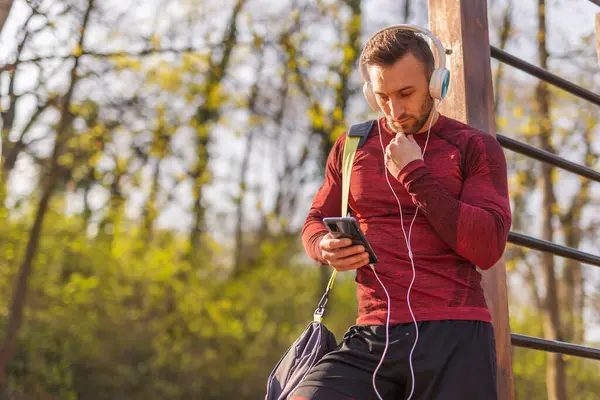 活跃的年轻人头戴耳机 带着健身包 用智能手机 在户外运动后输入短信 — 图库照片