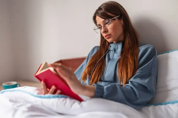 朝ベッドに座ってパジャマを着て 本を読んで自宅でリラックスする美しいペンギンな若い女性 — ストック写真
