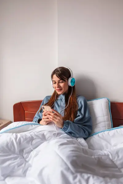 漂亮的年轻女子坐在床上 早上在家里休息 头戴耳机 用智能手机上网 — 图库照片