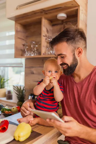 子供と一緒に家で楽しい時間を過ごしている父親 キッチンカウンターに座って 野菜を食べて漫画を見ている — ストック写真