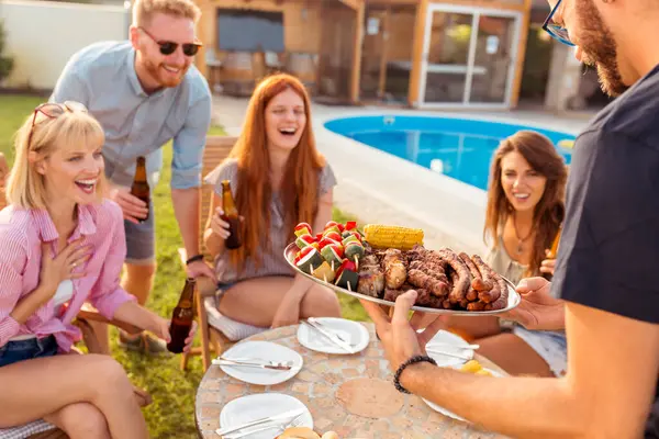 Group of cheerful young friends gathered around the table, drinking beer and having fun at a backyard poolside barbecue party, host bringing plate of grilled meat and vegetables