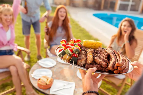 Group of cheerful young friends gathered around the table, drinking beer and having fun at a backyard poolside barbecue party, host bringing plate of grilled meat and vegetables