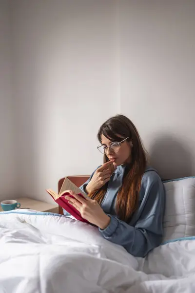 ベッドに座って本を読み 試験のために勉強するパジャマを着ている女性学生 — ストック写真