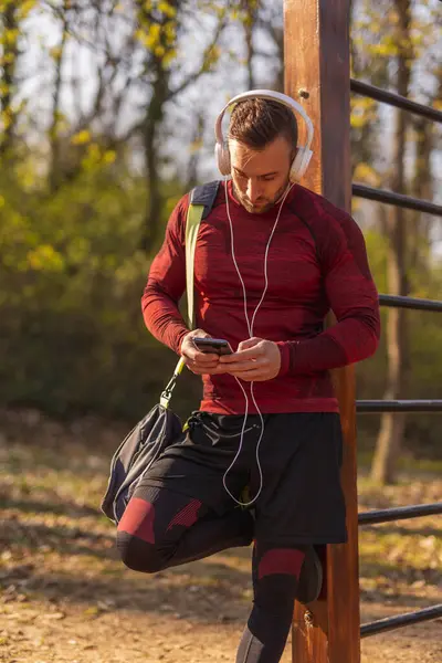 积极的年轻人头戴耳机 带着健身包 用智能手机 在户外运动前发短信 — 图库照片