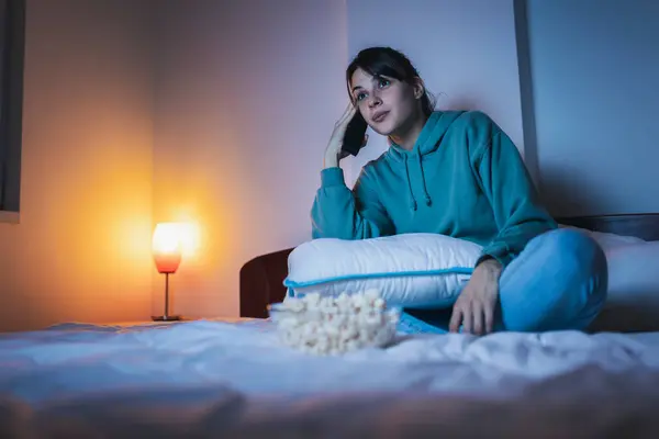 Beautiful young woman wearing pajamas sitting in bed at night, eating popcorn and watching a movie on TV