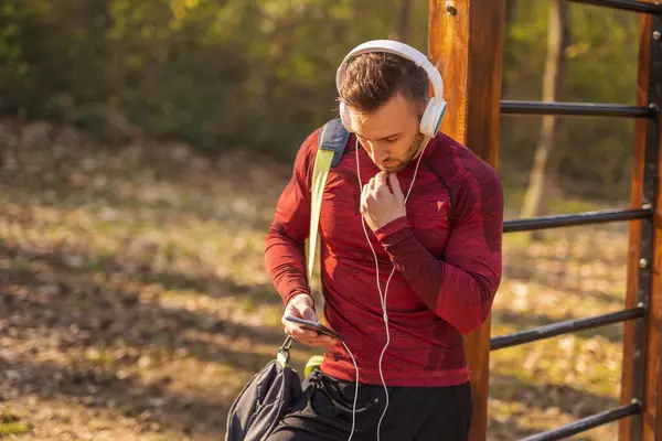 积极的年轻人头戴耳机 带着健身包 用智能手机 在户外运动前发短信 — 图库照片