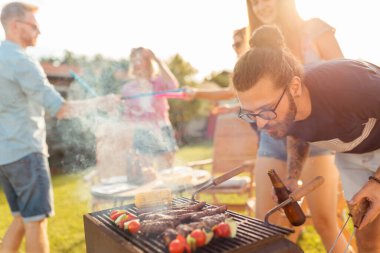 Bir grup arkadaş arka bahçede barbekü partisi veriyor, et pişiriyor, bira içiyor, badminton oynuyor ve güneşli bir yaz gününde eğleniyorlar.