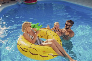 Genç ebeveynler yüzme havuzunda kızlarıyla oynarken eğleniyorlar, sıcak güneşli yaz günlerinin tadını çıkarıyorlar ve yaz tatilinde dinleniyorlar.